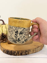 Load image into Gallery viewer, #001 - 16 oz. Leaf and acorn patterned speckled mug with burnt yellow rim and dark blue inside
